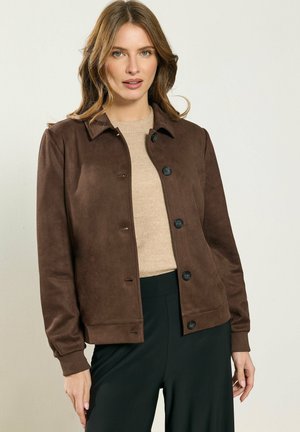 Donna che indossa una giacca marrone con bottoni sopra un maglione beige e pantaloni neri, in piedi contro uno sfondo chiaro e semplice.