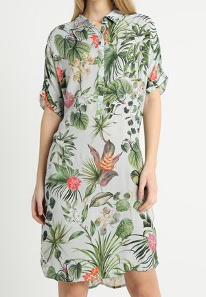 Vrouw draagt een korte mouw, knielange overhemdjurk met een groen en roze tropisch bloemenpatroon op een lichte achtergrond.