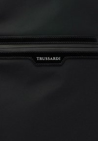 Černá taška z texturované tkaniny se značkovou černou koženou nášivkou s bílým embosovaným nápisem "TRUSSARDI" a detaily uzavřeného zipu.