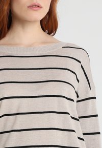 Femme aux cheveux roux portant un pull en tricot beige avec des rayures horizontales noires et un large col rond, sur un fond uni.