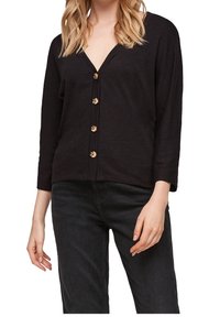 Femme portant un cardigan noir à col en V boutonné avec des manches trois-quarts et un jean foncé, visible du cou aux hanches.