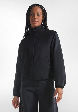 Junge Frau mit Zöpfen, die eine schwarze Jacke mit hohem Kragen und schwarze Hosen trägt, steht vor einem schlichten hellen Hintergrund.