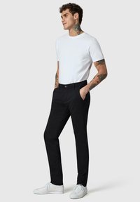 Homme avec des tatouages portant un t-shirt blanc à manches courtes, pantalon noir, baskets blanches, debout les mains dans les poches, regardant vers la droite.