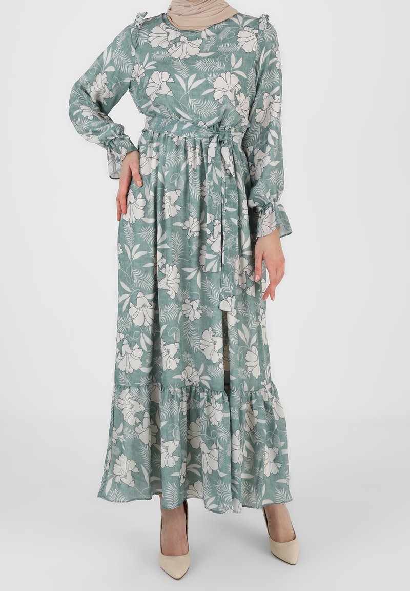 Femme portant une longue robe verte à fleurs avec une ceinture à la taille, des manches longues et des talons beiges, debout devant un fond uni.