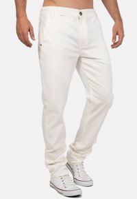 Man in slim-fit witte broek en witte sneakers, staand met de benen iets gekruist op een effen witte achtergrond.