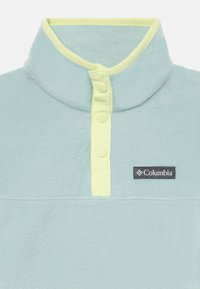 Lys blå fleece genser med høy krage, prydet med en gul kant og tre knapper; inkluderer et svart Columbia-logo på brystet.