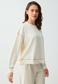Femme aux cheveux bruns mi-longs, portant un sweat-shirt oversized écru et un pantalon assorti, debout devant un fond clair uni.