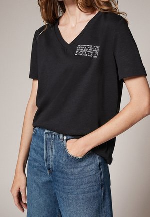 Czarny t-shirt z krótkim rękawem i dekoltem w serek o teksturowanym wykończeniu, z białym nadrukiem na lewej stronie klatki piersiowej, w połączeniu z niebieskimi jeansami o wysokim stanie.