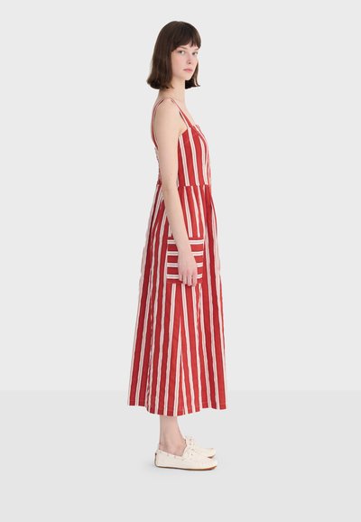 Femme debout de profil portant une robe maxi sans manches à rayures verticales rouges et blanches avec une poche, associée à des baskets blanches.