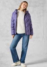 Lila puffjacka med huva, med dragkedja fram och elastiska ärmmuddar, bärs över en ljus tröja och kombineras med blå jeans och vita sneakers.