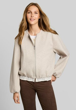Junge Frau mit langen Haaren trägt eine beige Reißverschlussjacke und braune Hose, steht mit einer Hand in der Hüfte vor einem grauen Hintergrund.
