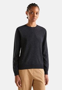 Kobieta z warkoczami, ubrana w ciemnoszary sweter z okrągłym dekoltem i beżowe spodnie, stojąca na jednolitym jasnym tle.