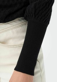 Svart strikket genser med teksturert mønster, med ribbestrikkede mansjetter og lett stoff, parret med lys denim.