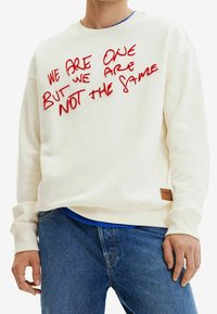 Sudadera color crema con texto bordado en rojo. Presenta un cuello redondo y puños acanalados. Parche de cuero notable en el dobladillo.