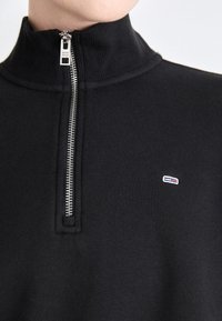 Maglione nero con zip alta, dotato di zipper argentato. Include un piccolo logo ricamato con accenti blu, rossi e bianchi. Texture liscia.