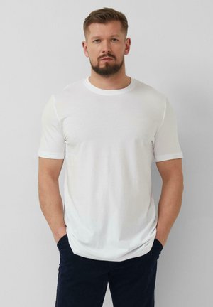 Man met baard draagt een effen wit T-shirt met korte mouwen en donkere broek, staat met handen in de zakken tegen een effen lichte achtergrond.