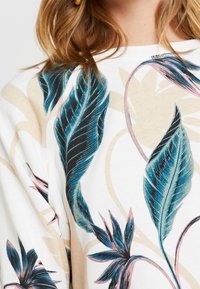 Sweat-shirt avec une base blanche ornée de feuilles multicolores dans des tons de bleu, rose et vert, sur un motif floral beige subtil.