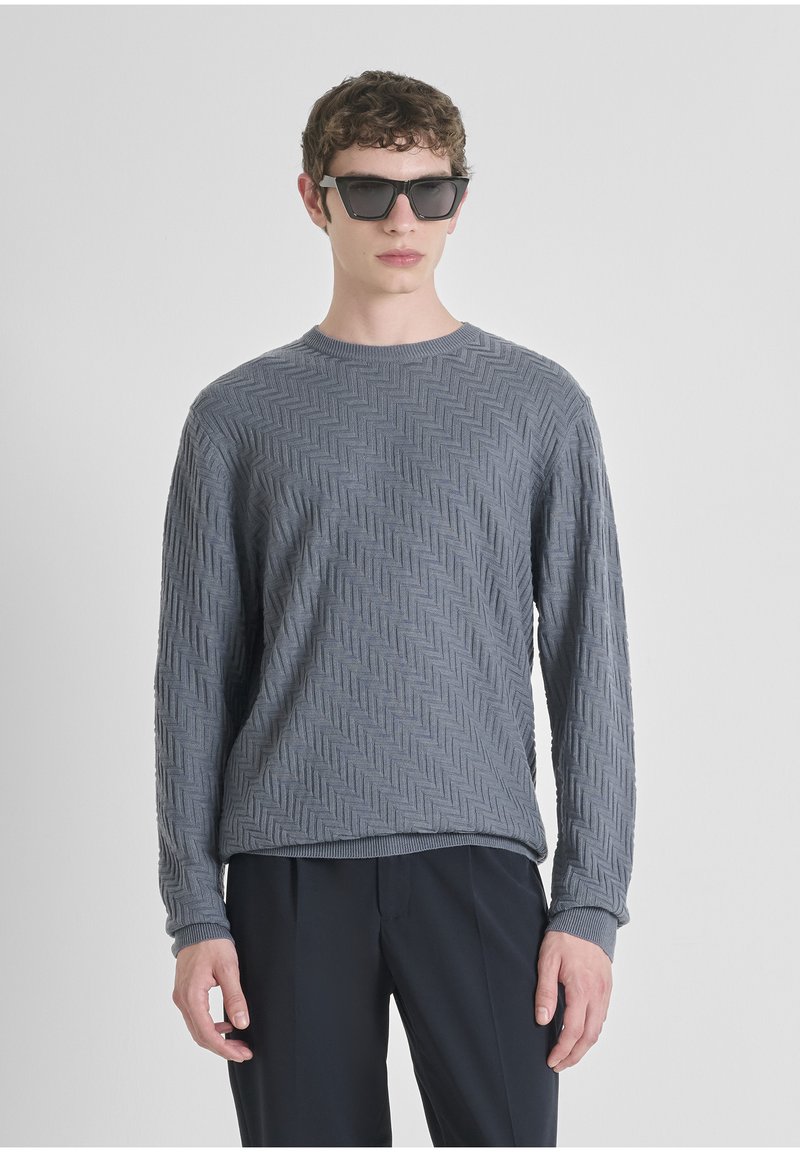 Maglione grigio a maniche lunghe con un motivo herringbone testurizzato, scollatura a girocollo e polsini a coste, abbinato a occhiali da sole neri angolari.