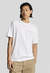 Hvid kortærmet T-shirt i bomuld med grafisk print på venstre bryst. Runde halsudskæring og afslappet pasform, parret med beige shorts.