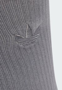 Šedá žebrovaná ponožka s vyšívaným logem zobrazujícím tři listy a vodorovné pruhy poblíž kotníku. Měkká textura a neformální design.