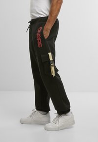 Mann iført svarte cargo joggebukser med rødt "ONCRS"-trykk og beige sidebuckle, hvite sokker og hvite Nike sneakers, som står mot en grå bakgrunn.