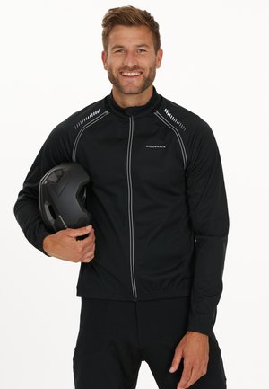 Veste de cyclisme noire avec accents réfléchissants, fermeture éclair à l'avant, poignets élastiques, matériau léger. Portée avec un casque noir tenu dans une main.