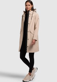 Beige vattentät jacka med huva, tryckknappar och två sidofickor. Bärs över en svart topp och leggings, tillsammans med ljusa sneakers.