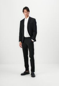 Costume noir avec un design ajusté, fermeture à un bouton, associé à une chemise blanche. Le mannequin porte des chaussures noires, se tenant devant un fond neutre.