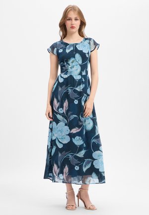Jeune femme debout portant une robe longue bleu marine à fleurs avec des manches courtes et des sandales beiges à talons hauts sur un fond blanc.