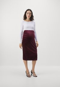 Oblačilo za materinstvo z burgundskim žametnim krilom in prilegajočim se belim dolgim rokavom. Oblikovanje vključuje raztegljiv pas in eleganten silhuet.