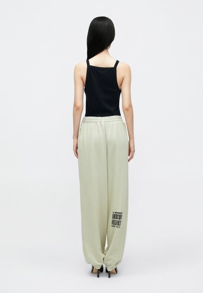 Czarny top na ramiączkach, jasnoróżowe dresy z elastycznymi ściągaczami na nogawkach i nadrukiem graficznym na lewej nodze, noszone z czarnymi obcasami. Gładki materiał i dopasowany krój.