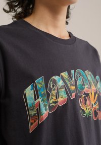 Melna kokvilnas T-kreklā ar krāsainu "Havana" grafiku, kurā ir tropu attēli un rozā un zilas akcenti izteiksmīgā, stilizētā fontā.