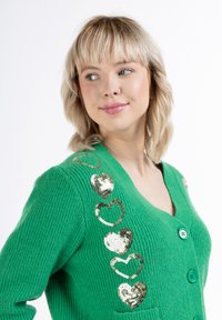 Cardigan verde lavorato a maglia con scollo a V, cinque bottoni e dettagli a forma di cuore con paillettes dorate lungo il lato sinistro. Tessuto texture e tasche incluse.