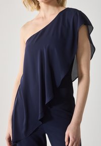 Tamno plava bluza s jednim ramenom s preklapajućim materijalom, koja ima mekan draper i lepršavi rukav, izrađena od laganog, glatkog materijala.