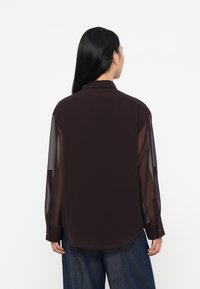 Mørk burgunderbluse med gjennomsiktige ermer, klassisk krage og knappede mansjetter. Glatt tekstur, lengre bakhem og rett snittdesign.