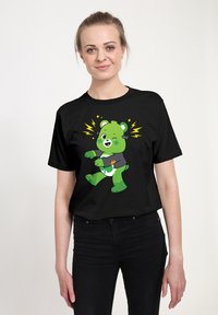 Camiseta negra con un oso verde de dibujos animados que tiene una expresión guiñando un ojo, vistiendo una camiseta gris, rodeado de rayos amarillos.