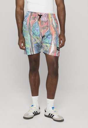 Man draagt meerkleurige korte broek met patroon, witte tanktop, witte sokken en witte sneakers met zwarte strepen, staand tegen een effen achtergrond.