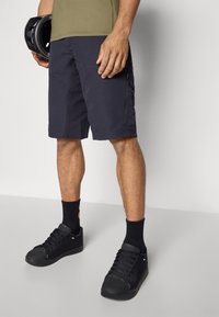 Shorts de sport noirs avec une finition texturée, arrivant au genou, portés avec des baskets noires et des chaussettes noires. La silhouette tient un casque noir.