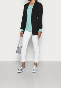 Schwarzer maßgeschneiderter Blazer über einem türkisfarbenen Hemd, kombiniert mit weißen Jeans, die bis zu den Knöcheln hochgerollt sind, und silbernen Turnschuhen. Sie trägt eine kleine graue Handtasche.