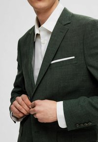 Tumši zaļš bleizeris ar teksturētu apdari, ar baltu kabatas lakatiņu, iegrieztiem atlokiem un pogātiem aproces pār baltu krekls.