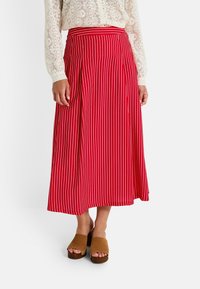 Falda midi a rayas rojas con una cintura ajustada, tela fluida y un diseño plisado. Combinada con sandalias de plataforma marrones.