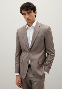 Completo marrone chiaro con finitura testurizzata, caratterizzato da un design a un bottone, due bottoni, rever a punta e tasche frontali. Camicia bianca sotto.