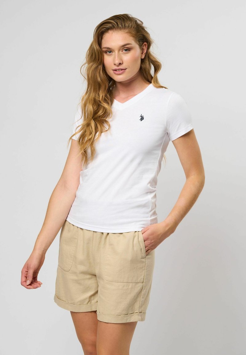 Witte V-hals t-shirt met korte mouwen, met een klein zwart logo op de linkerborst; gecombineerd met tan linnen shorts met zakken.