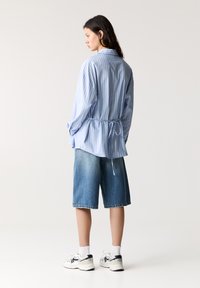 Ljusblå randig skjorta med knytning i midjan, kombinerad med denimshorts i Bermuda-stil och vita sneakers med svarta detaljer. Slät tygtextur.