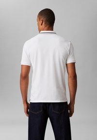 White polo shirt with short sleeves, featuring a ribbed collar and subtle horizontal stripes. Worn with dark blue denim jeans.