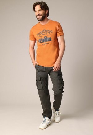 Oranje korte mouwen graphic t-shirt met blauw vrachtwagenontwerp, gecombineerd met donkere cargo-broek met zijzakken en witte sneakers.