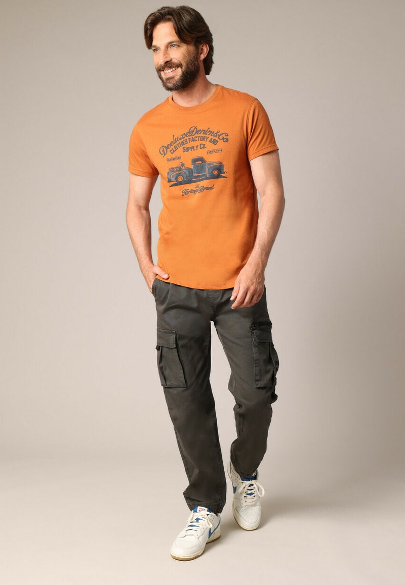 Oranje korte mouwen graphic t-shirt met blauw vrachtwagenontwerp, gecombineerd met donkere cargo-broek met zijzakken en witte sneakers.