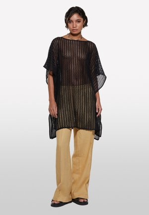 Tunique noire, légère, en tricot ouvert avec une coupe ample et des manches larges, associée à un pantalon beige à jambes larges et des sandales à plateforme noires.