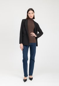 Blazer nero con bottoni doppi su un dolcevita marrone, abbinato a jeans blu scuro e décolleté nere a punta. Sfondo semplice.