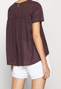 Bordeaux rode blouse met korte mouwen en witte polka dots, met een gerimpeld ontwerp en knoopdetails op de rug, gecombineerd met witte shorts.